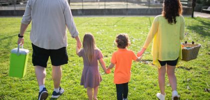 A family of four walking hand in hand through a sunny park carrying picnic supplies.